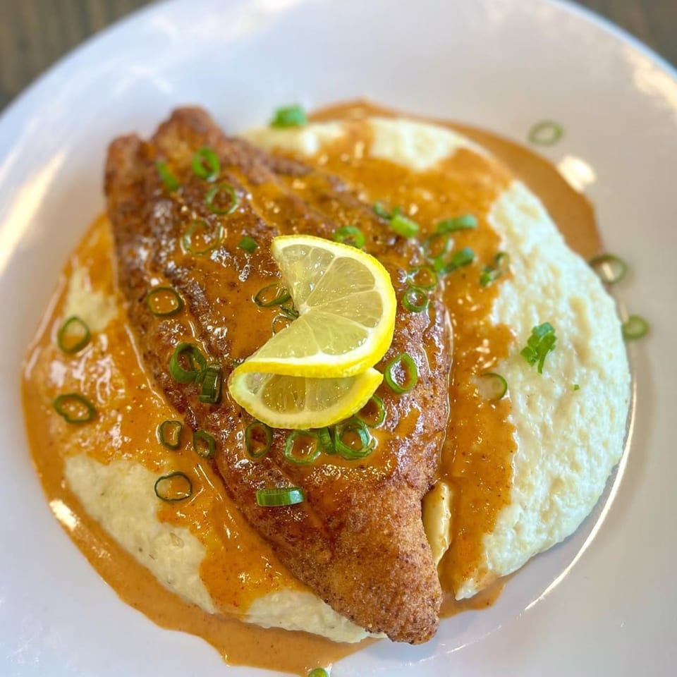 Crispy fried fish with creamy grits, a rich orange sauce, green onions, and lemon slices.