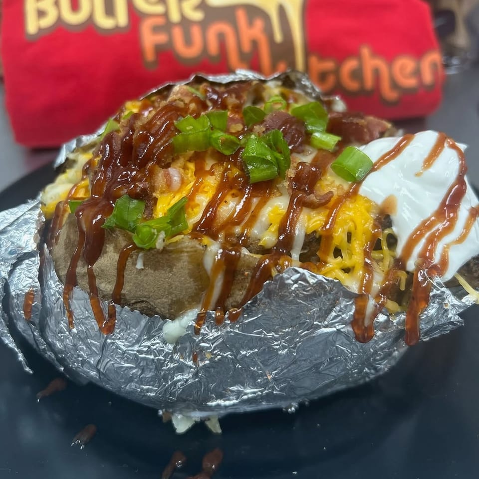 A loaded baked potato from Butter Funk Kitchen, topped with cheese, bacon, green onions, BBQ sauce, and sour cream.