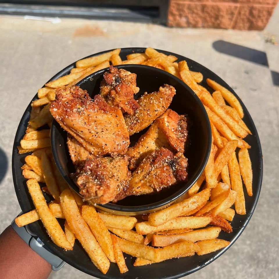 A black plate holds a bowl of seasoned fried chicken wings surrounded by golden french fries.